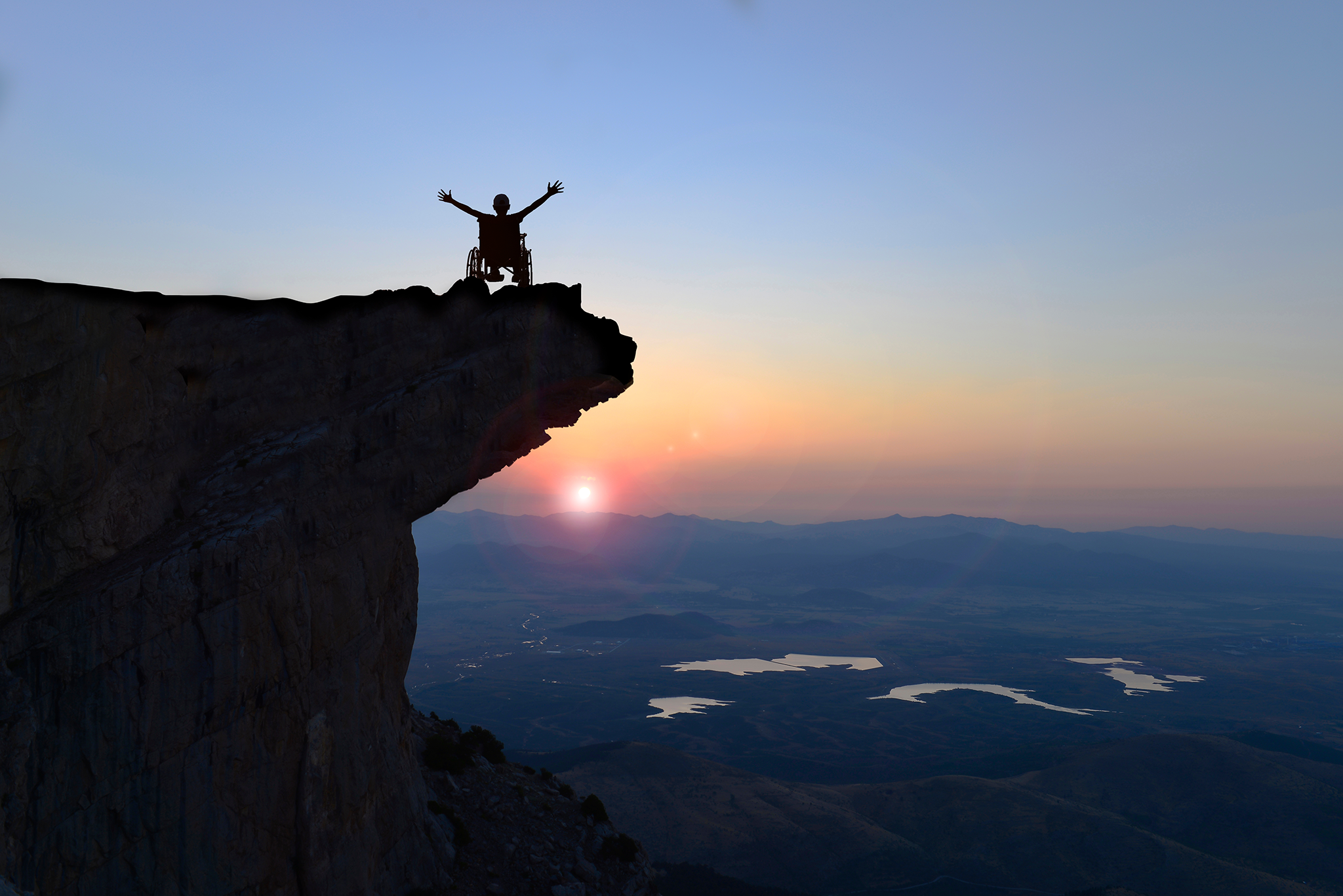 Une personne en fauteuil roulant est au-dessus d’une montagne et lève les bras en signe de victoire face au coucher du soleil. Photo.